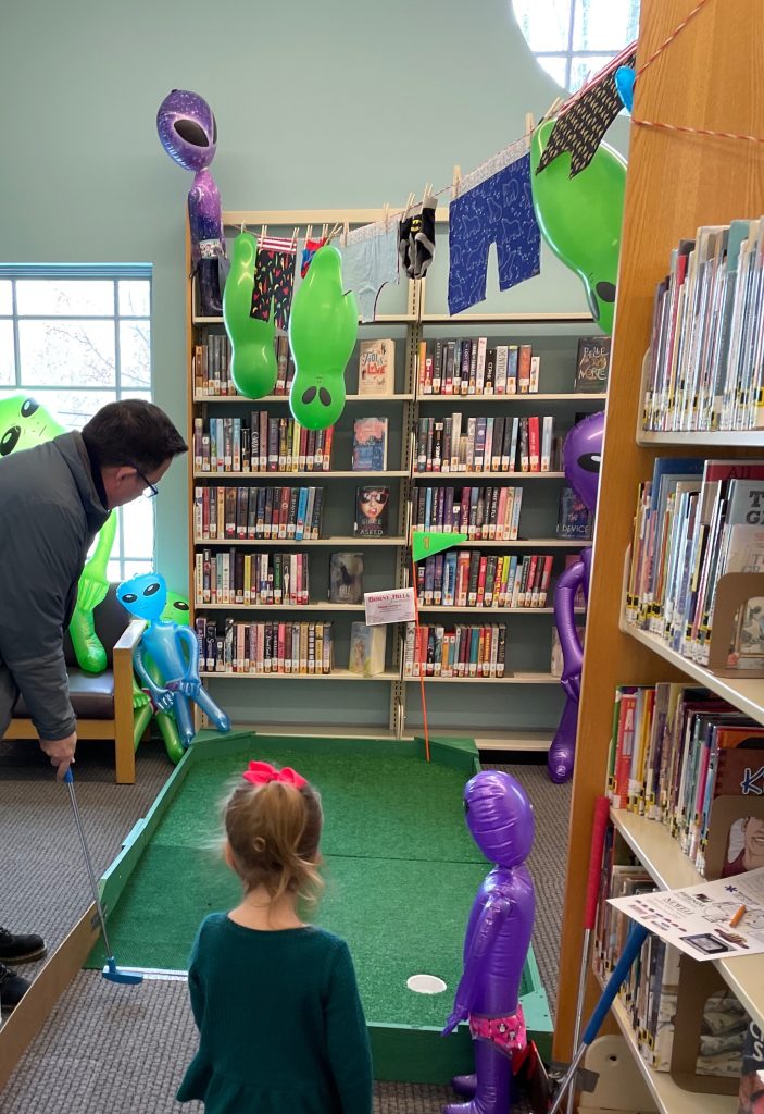 a man plays mini-golf in the aisles of the library as a young child looks on.